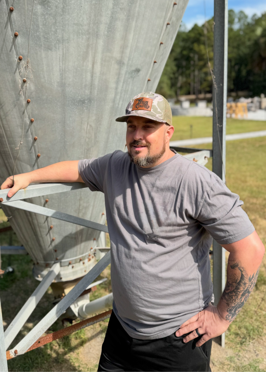 Man wearing a cap and gray t-shirt leaning against a large metal structure outdoors.
